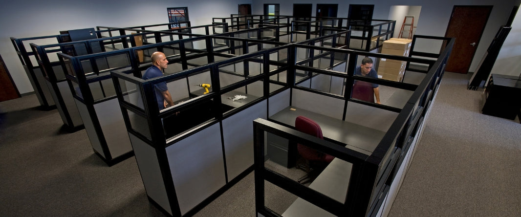 An office space with multiple cubicles under construction. Workers are seen installing partitions and furniture