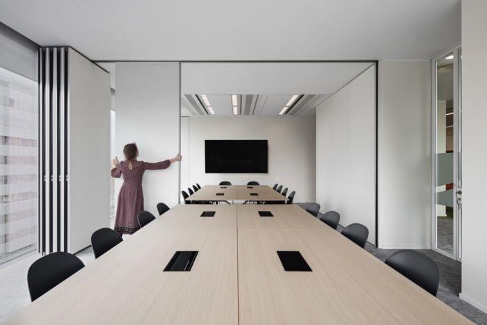 Modern meeting room with a movable partition wall being adjusted by a person to divide the space. The room features minimalist furniture, including long tables and black chairs, with a screen mounted on the wall.