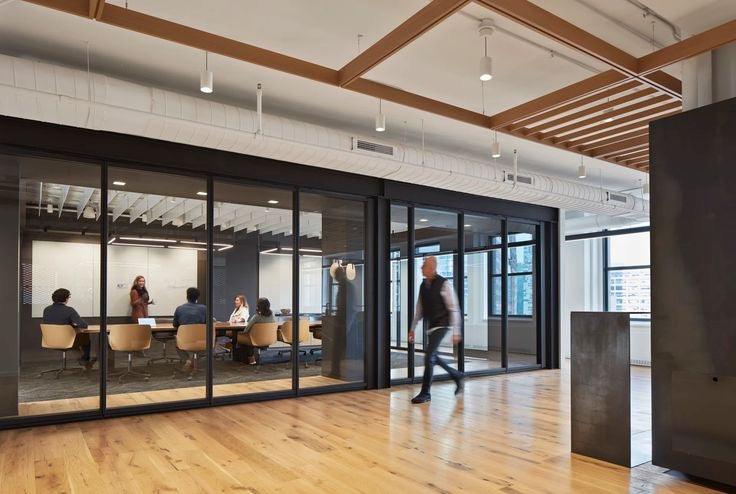 Modern office environment with a glass foldable partition wall separating a conference room. The space features wooden floors, exposed ceiling beams, and a contemporary design, with people engaged in a meeting inside the partitioned area.