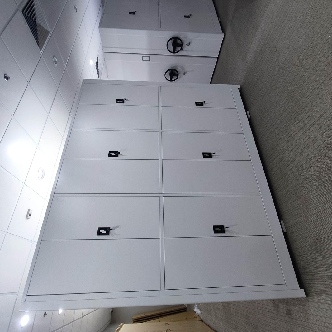 A modern office storage room with white large file compactor, lockable compartments and a manual wheel for easy movement. The space is well-lit with recessed ceiling lights.