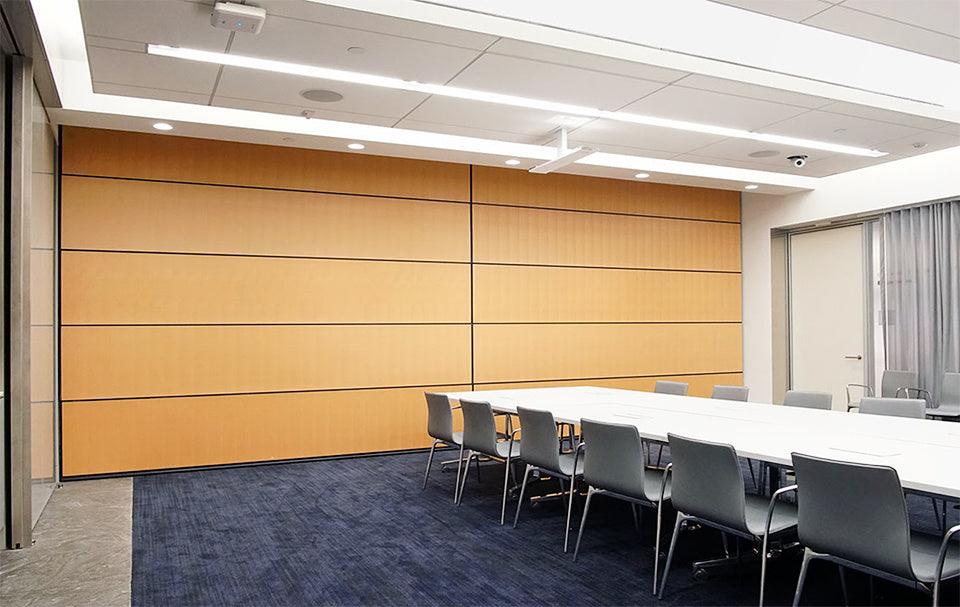 Conference room with acoustic movable partition wall, featuring wood finish, dividing the space. The room is set up with a long meeting table and modern chairs, illuminated by ceiling lights. 