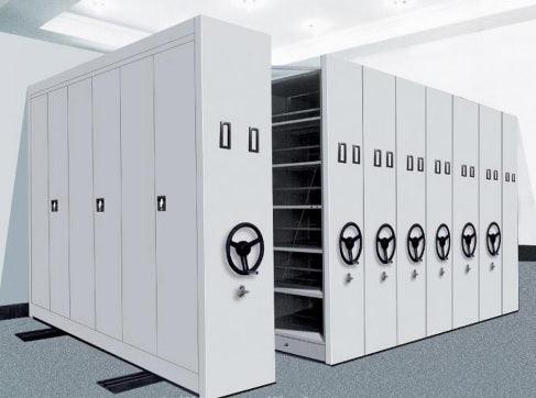 File compactor system with multiple mobile units on tracks, equipped with crank handles for easy access. The compactors have lockable doors and adjustable shelves, designed for high-density file storage in an organized, space-efficient manner.