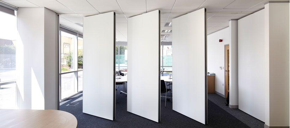 Bright office space featuring a collapsible room divider in a white finish, partially open to divide the room. Large windows provide natural light, with the divider offering flexible space management.