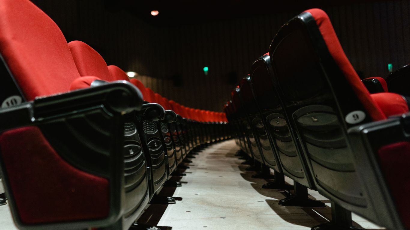 A side view of a row of red theater seats, with the seats curving away to create a sense of depth. The seats have black armrests and appear to be upholstered in a plush fabric.