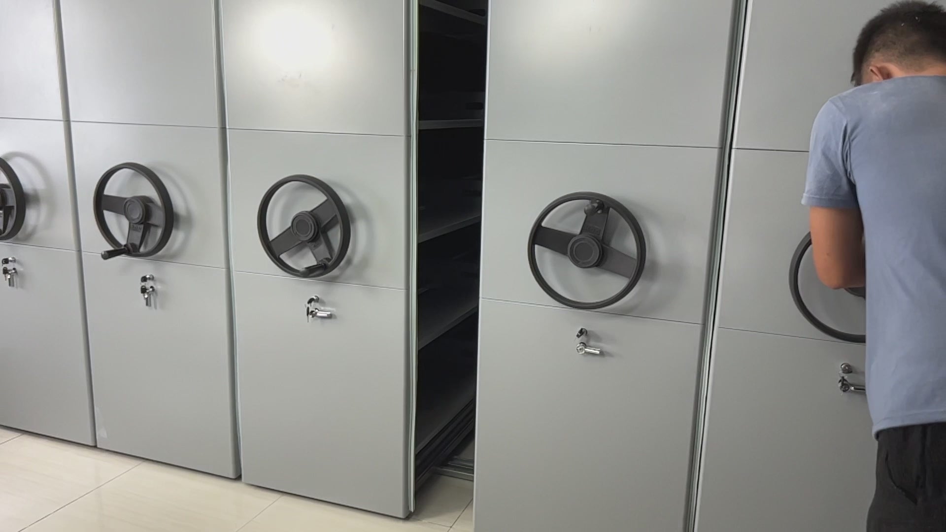 A man operating a file compactor system, turning the black rotating handle on a gray metal unit with lockable compartments, designed for organized file storage.